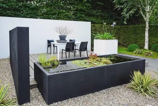 Black raised pond with water plants set in a gravel courtyard beside a white wall and timber deck
