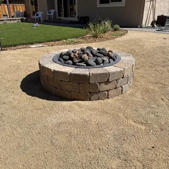 Round stacked-stone fire pit with lava rock set on a compacted gravel base beside a lawn