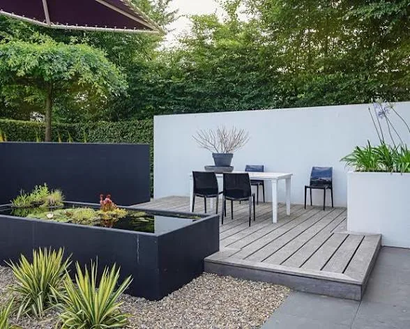 Timber deck with outdoor dining table set beside a raised black pond, white walls and clipped hedging