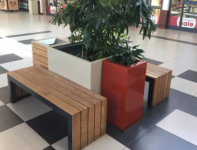 Planter box with tall foliage flanked by slatted timber benches on a tiled shopping centre floor