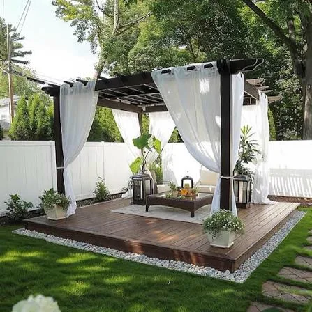 Low timber deck with a dark pergola, white drop curtains, lounge seating and a pebble border on a lawn