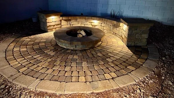 Circular stone-paved fire-pit area with a curved seating wall and pillar lighting at night
