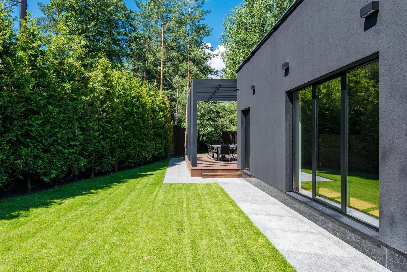 Striped manicured lawn beside a dark modern house with a slatted pergola over a timber dining deck