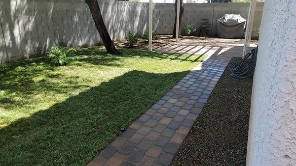 Brick paver pathway running beside a shaded lawn and boundary wall with a gravel strip along the house
