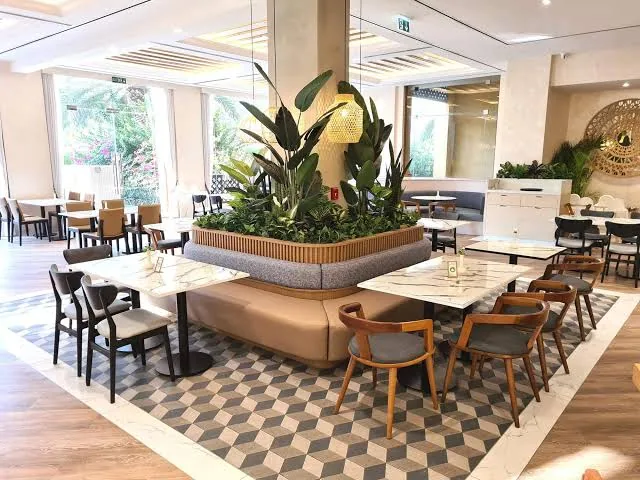 Timber-clad indoor planter box with tall green foliage set between marble dining tables in a restaurant